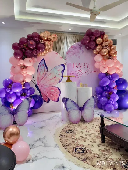 An elegant butterfly theme for a baby shower, set up in a home. This design uses two curved backdrops, large butterfly cutouts, and a rich color palette of purple, pink, and rose gold balloons to create a stunning focal point.