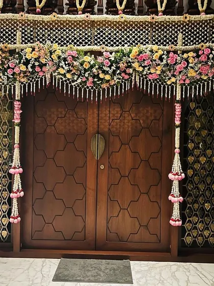 This pooja room door was decorated with a beautiful fusion of traditional flowers. A net of jasmine flowers, or 'mallepuvu jada', is framed by intricate floral pillars and a colorful toran for a housewarming event.