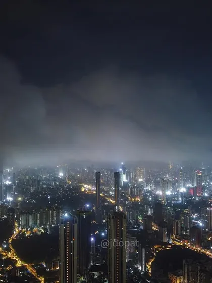 A wide drone shot of Mumbai at night, showing a bank of monsoon clouds rolling over the illuminated cityscape.