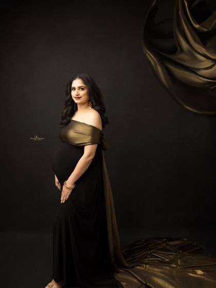 A simple yet stunning portrait against a black backdrop. The off-shoulder black and gold gown drapes beautifully, highlighting the mother-to-be's elegant silhouette.