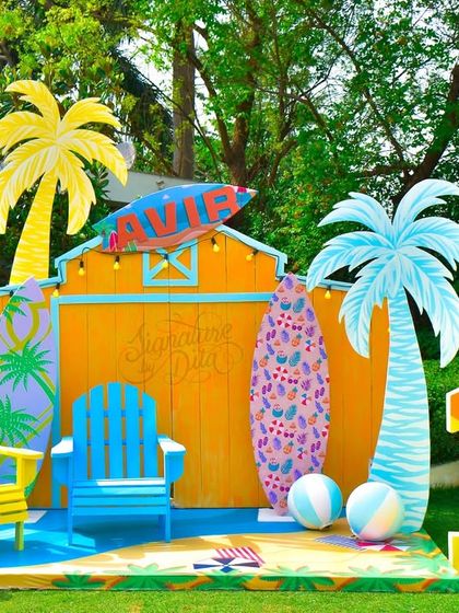 A vibrant photo booth for a beach-themed 10th birthday. The setup includes custom-painted surfboards, palm tree props, and a large number '10', all designed for a fun, sunny outdoor celebration.