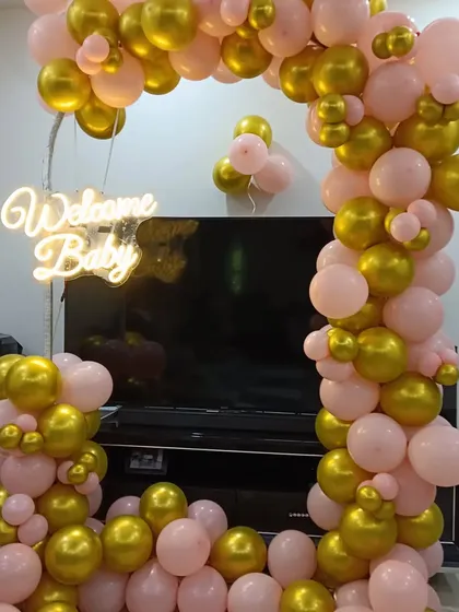 An elegant 'Welcome Baby' balloon arch in pink and gold, designed to frame a TV or fireplace area in a home. The setup is highlighted by a warm white neon sign, making it a beautiful focal point.