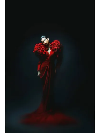 A dramatic, low-key portrait of Athena in a red gown with large floral shoulders. The single light source from above creates a theatrical and mysterious mood.