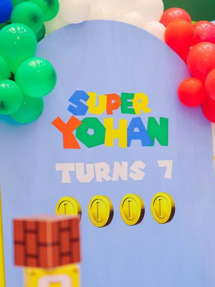 A "Super Yohan Turns 7" sign with gold coin graphics, set against a backdrop of game-inspired scenery and colorful balloons.