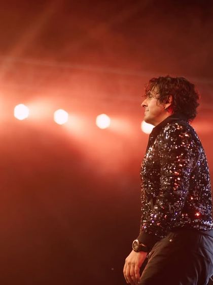 A profile shot of Sonu Nigam against a backdrop of red lights. The focus is on his silhouette and the energy radiating from the stage.