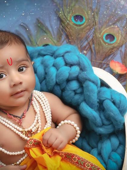 A close-up of a baby as little Krishna, with beautiful peacock feathers in the background. The focus is on his innocent expression and traditional attire.