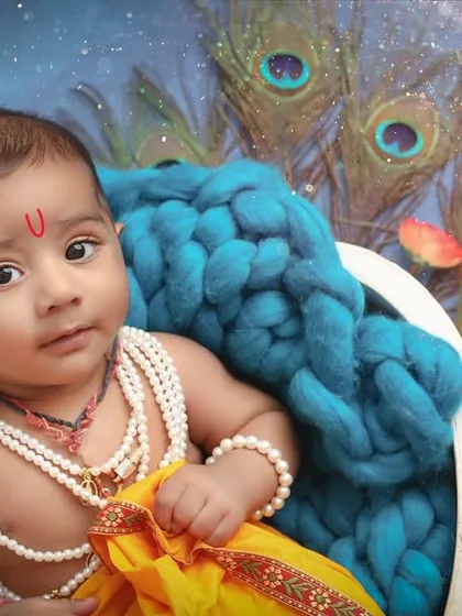 A close-up of a baby as little Krishna, with beautiful peacock feathers in the background. The focus is on his innocent expression and traditional attire.