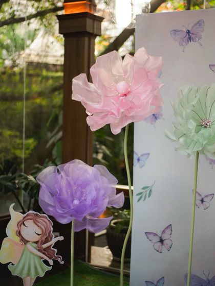 Giant, ethereal paper flowers in pastel pink, purple, and green stand against a butterfly-print backdrop, adding a touch of whimsical beauty to our fairy parties.