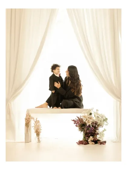 A mother and her young son, dressed in matching black suits, share a look of pure connection in my all-white studio. The minimalist setting, framed by soft curtains, strips away everything but the love and joy between them, creating a modern, artful family portrait.