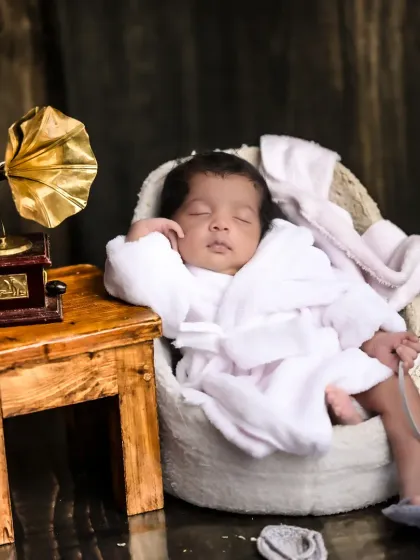 A classic newborn portrait featuring a baby in a cozy robe, looking relaxed and content in a miniature armchair next to a vintage gramophone.