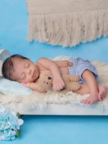 A baby boy in blue shorts sleeping on a tiny bed with his teddy bear.