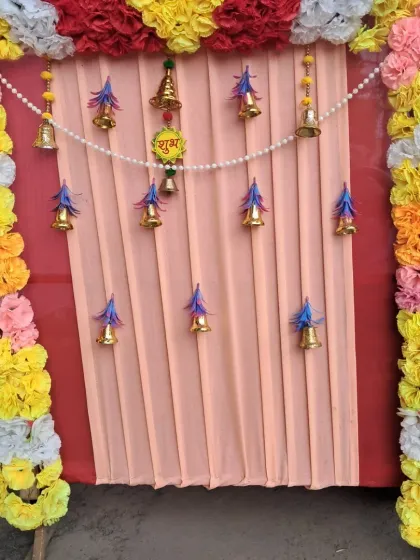 A colorful backdrop using yellow, pink, and white artificial flowers. The design includes a string of pearls and multiple hanging bells against a pleated peach fabric background for a traditional festive look.