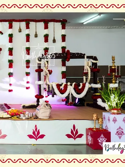 An elegant and traditional Namakaran (naming ceremony) setup. The centerpiece is a beautifully decorated wooden cradle, or 'jhoola', adorned with garlands of red and white flowers, set against a backdrop of floral hangings and brass bells.