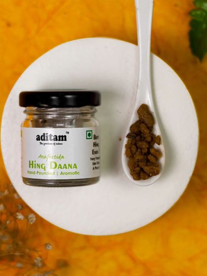 A small jar of Hing Daana (asafoetida) is presented on a simple white dish. The warm, yellow background and minimalist styling keep the focus on the unique texture and color of the spice.