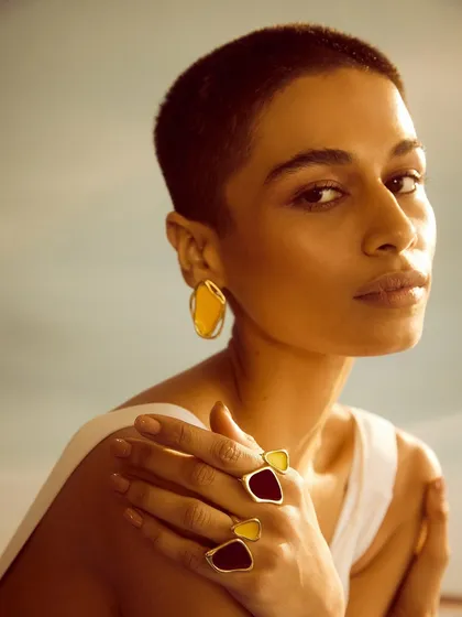 A warm, sun-kissed portrait of the model. The close-up allows the viewer to appreciate the craftsmanship of the enamel earrings and rings.