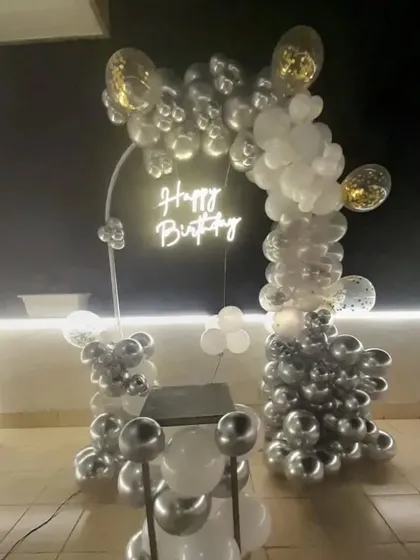 A sleek outdoor birthday setup at night with a silver and white balloon arch and a 'Happy Birthday' neon sign.