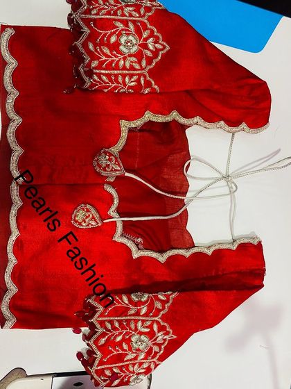 A bright red blouse with silver Zari work. The scalloped edges on the sleeves and back give it a modern look, and the tie-ups have heart-shaped details.