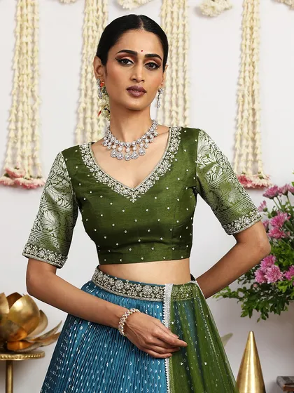 A close-up of the olive green embroidered blouse, featuring brocade sleeves and delicate pearl detailing. It is paired with a teal blue Bandhani lehenga.