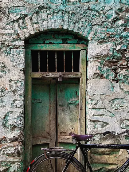 A weathered green door is set within a rough, turquoise-painted stone wall. A bicycle parked in front adds a touch of daily life to this scene rich with texture and history.