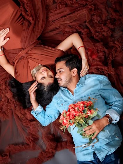 A creative top-down view of the couple lying on the rich, brown fabric of the gown. The partner holds a bouquet, adding a romantic, artistic touch.