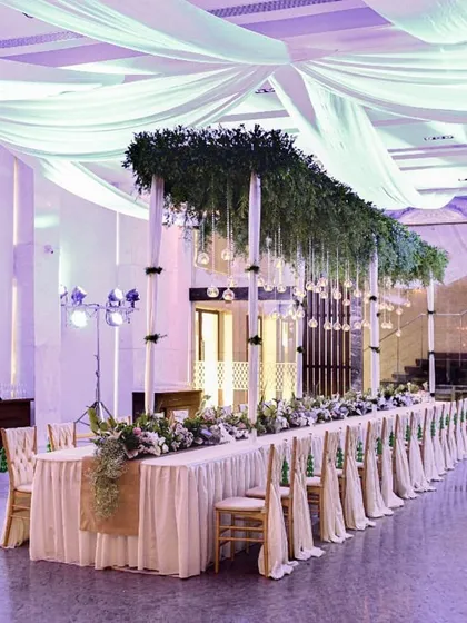 A grand dining hall with a long, elegantly decorated head table. The lush greenery and flowing drapes create a sophisticated and dramatic setting for a large celebration.