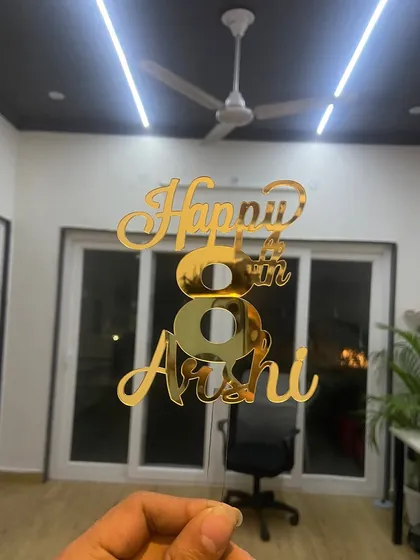The "Happy 8th Arshi" topper held against an indoor background, showing how its reflective surface shines even in regular lighting.