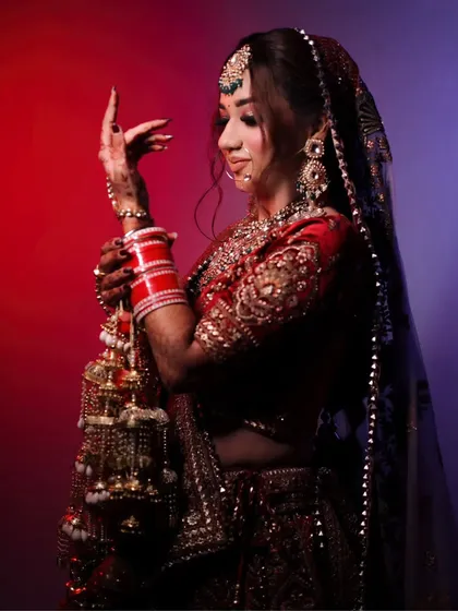 A stunning full-length bridal portrait showcasing the bride's complete look. The pose highlights her traditional red lehenga, the intricate kalire, and her graceful posture against a dramatic, colorful background.
