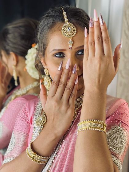 A beautiful shot of a bride showcasing her long, elegant nail extensions. The design is a soft, shimmery pink that perfectly matches her traditional pink and gold saree.