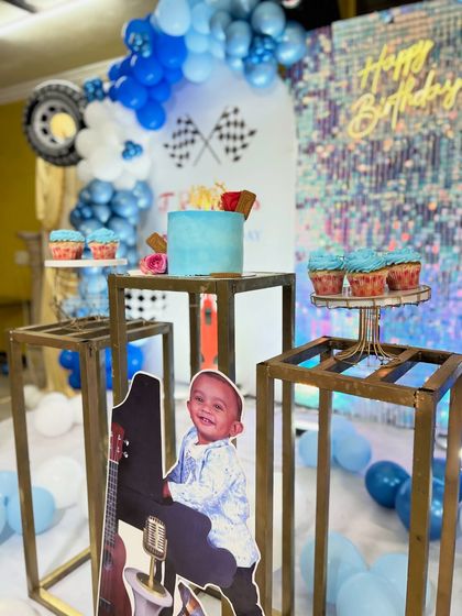 The complete dessert station for the car-themed party, featuring a custom cake and cupcakes on themed plinths, set against the shimmering backdrop and balloon arch.