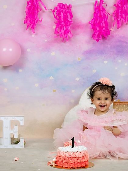 A classic first birthday portrait. The little girl in her pink tulle dress is all smiles, sitting beside her cake and the light-up "ONE" sign.