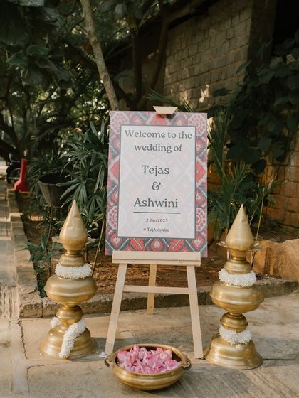 A welcome sign for the wedding of Tejas & Ashwini, flanked by traditional brass lamps.