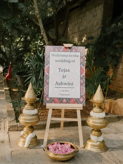 A welcome sign for the wedding of Tejas & Ashwini, flanked by traditional brass lamps.