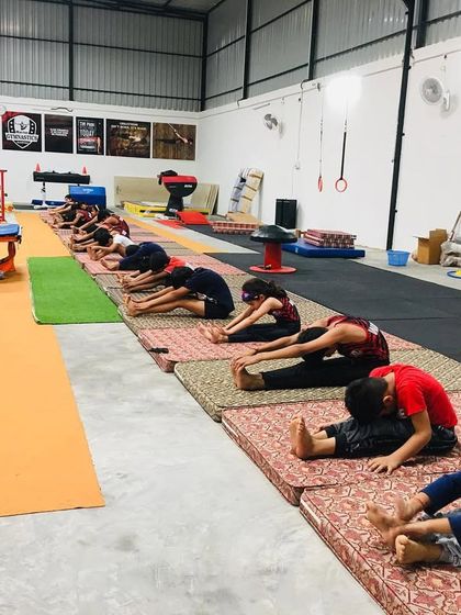 A coach supervises a group of young gymnasts during their seated forward fold stretches. We emphasize proper technique from a young age to develop lifelong mobility.