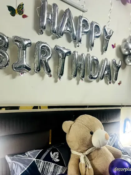 A detailed shot of the teddy bear and 'Happy Birthday' foil balloons. The soft lighting from the bedside lamps adds to the cozy and celebratory atmosphere of the room.