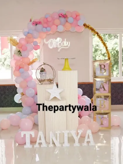 A lovely baby shower decoration with a pastel pink and blue balloon arch. The setup includes "BABY" blocks and a small pedestal, perfect for a home celebration.