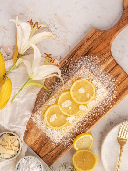 A bright and airy shot of a lemon and almond loaf cake. The styling with fresh flowers and lemon slices creates a light, fresh feeling, perfect for a spring or summer menu. The powdered sugar adds a touch of delicate sweetness.