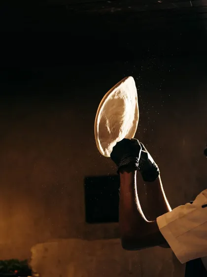 A chef tosses a whole wheat pizza base into the air. This isn't just for show; it helps to evenly stretch the dough to the perfect thinness without compressing the edges.
