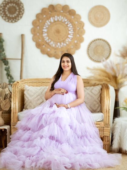A smiling mom-to-be seated on a cane sofa, the lavender gown spreading out beautifully around her.