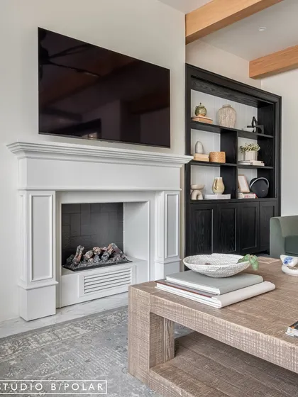The feature wall of the living room is anchored by a classic white fireplace, creating a traditional focal point. Flanked by built-in display units, the design feels balanced, grand, and inviting.