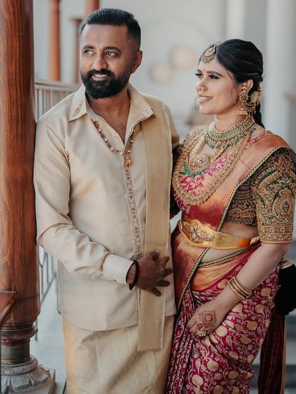 A couple posing by a pillar, dressed in our bespoke creations. Her rich brocade saree with a heavily embroidered green blouse is perfectly balanced by his simple yet elegant cream silk shirt and dhoti.