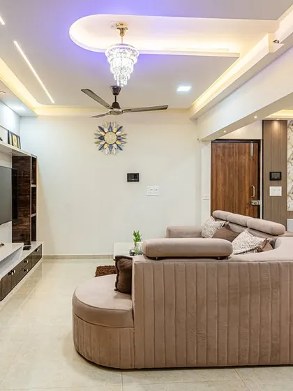 This living room features a large, comfortable sectional sofa perfect for family time. The false ceiling design with cove lighting and a central chandelier adds a sense of grandeur and warmth to the space.