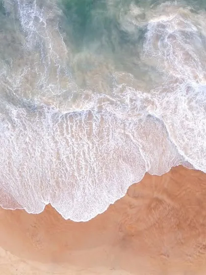 A top-down aerial photograph of the Goa coastline, showing the beautiful contrast between the turquoise water, white seafoam, and the warm-toned sandy beach.