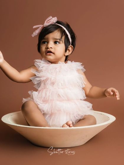 A minimalist portrait of a baby girl in a pink ruffled romper, sitting in a simple bowl against a brown backdrop.