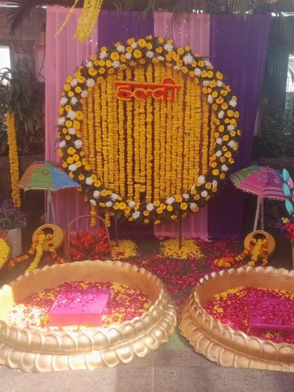 A grand Haldi setup with a circular marigold backdrop and two large brass urulis filled with flower petals. The purple and pink drapes add a touch of contrast to the traditional yellow theme.