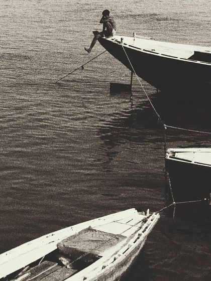 This black and white photograph captures a sense of solitude and contemplation, as a lone figure sits on the bow of a boat in Varanasi. The minimalist composition and the vastness of the water emphasize the feeling of being in one's own world.