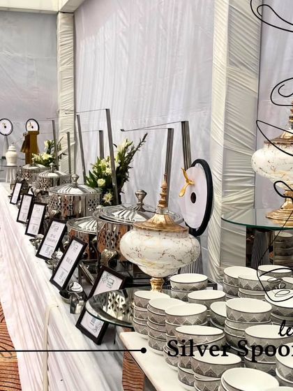 A sophisticated buffet line featuring a mix of modern silver serveware and decorative marble urns. The clean, white theme with floral accents is perfect for corporate events and formal gatherings.