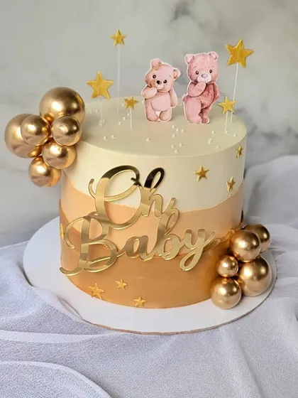 An elegant baby shower cake in a neutral color palette. The design features shades of beige and gold, with cute teddy bear toppers and a gold "Oh Baby" sign.