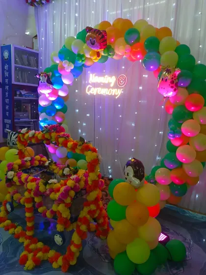 A jungle-themed naming ceremony with a beautifully decorated cradle (palna) covered in marigold flowers. The backdrop is a vibrant green and yellow balloon circle with a custom neon sign.