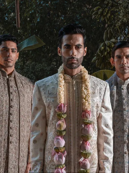 A powerful portrait of a groom flanked by his groomsmen. The focus is on the groom's majestic presence, enhanced by his finely detailed sherwani and a traditional lotus garland.