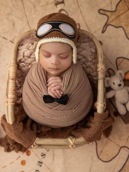 Ready for takeoff. This little aviator is swaddled and sleeping in a basket, wearing a pilot's cap and surrounded by a toy plane and teddy bear on a vintage map background.